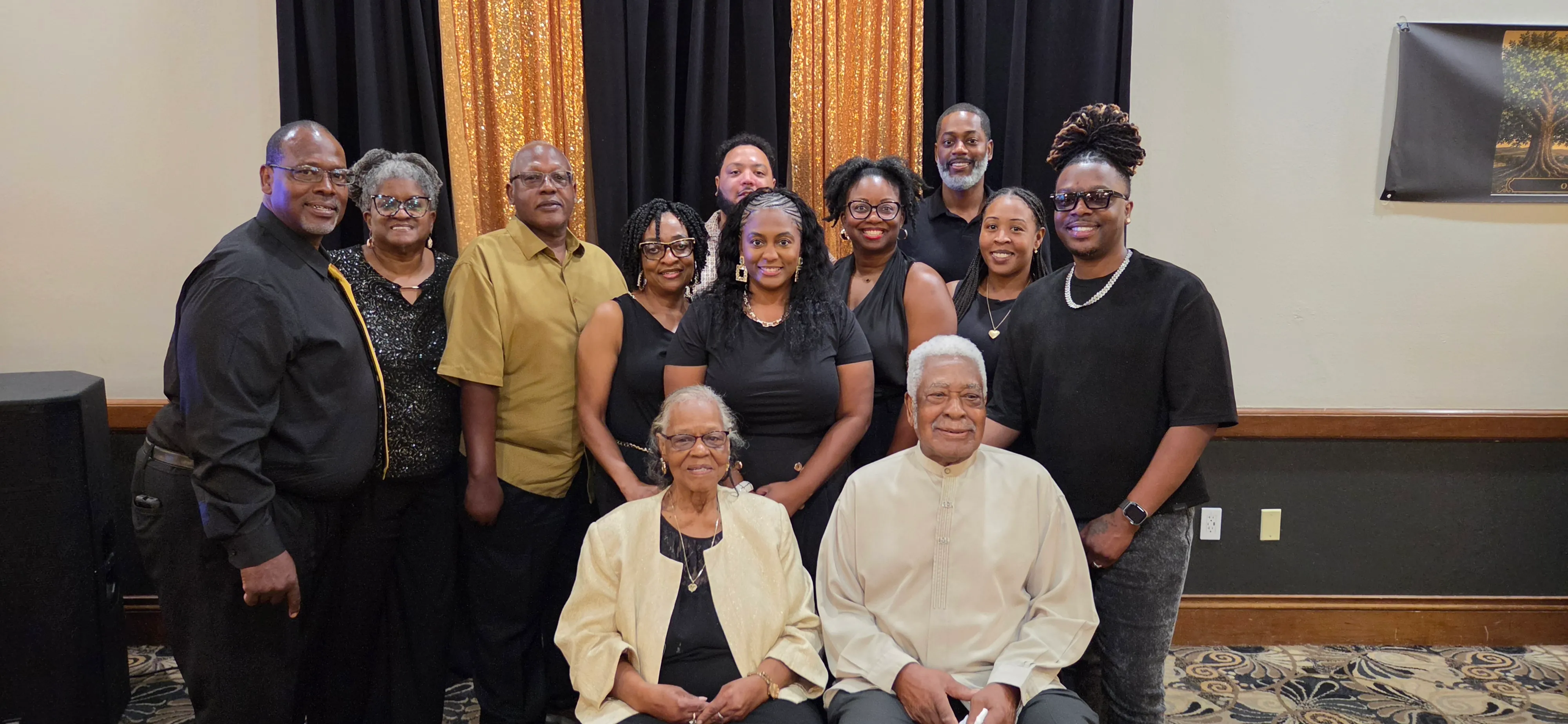 A large group dressed in black and gold posing in front of black and gold curtains, with two elders seated in front.