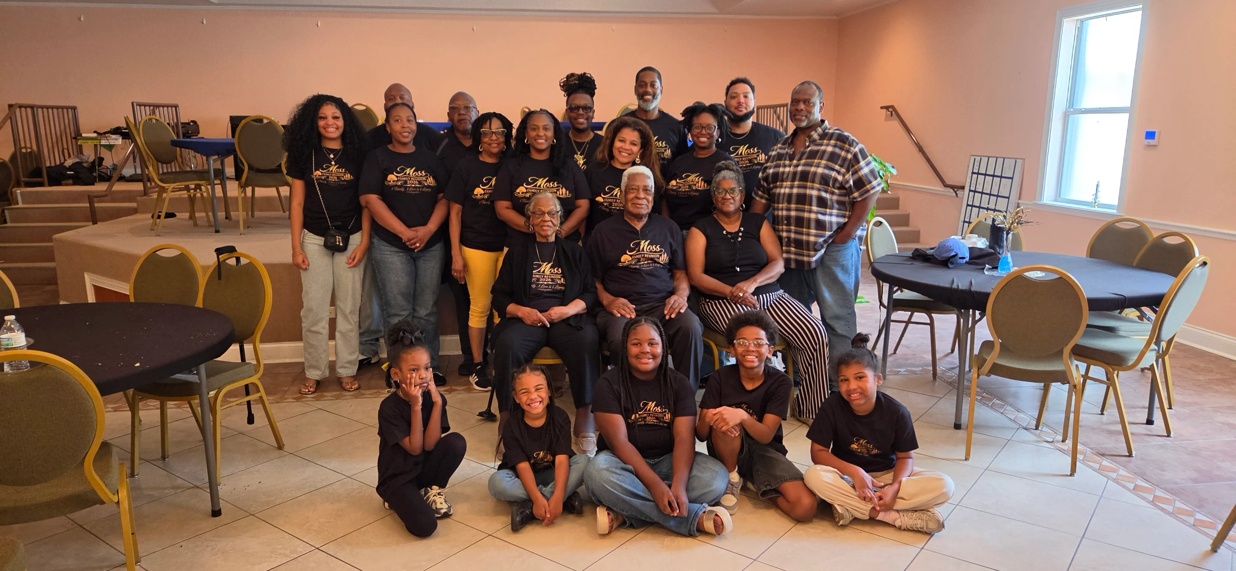 Family in black reunion shirts gathered in a hall. Adults stand in the back and children sit on the floor in front.